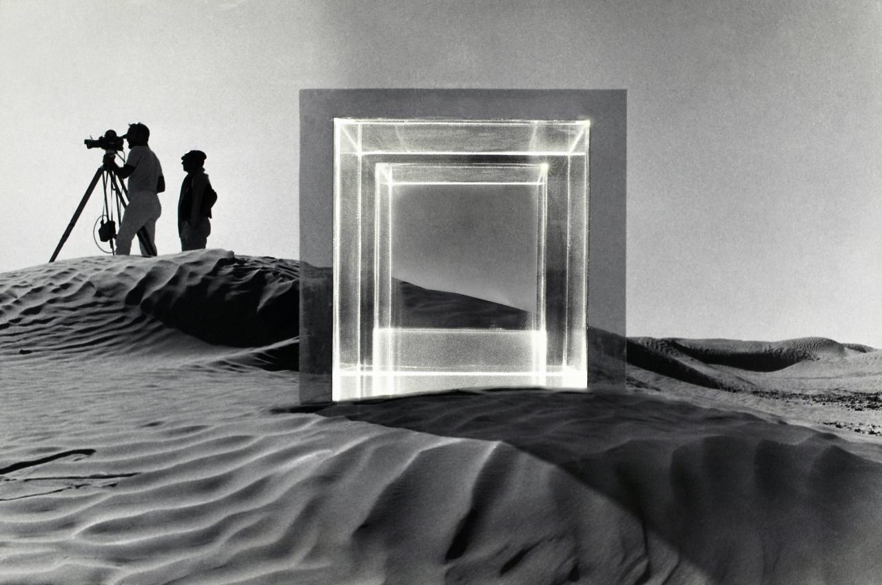 In a desert landscape around 1968, filming of the movie "Tele-Mack" took place. In the foreground is a large square glass sculpture and in the background are two men with a film camera. The picture is black and white.