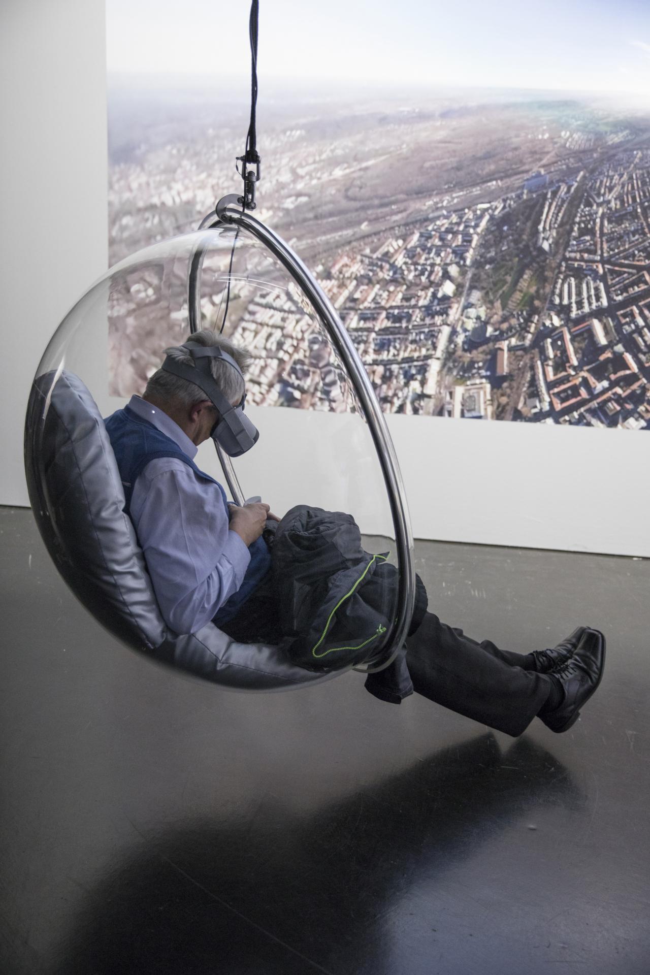 A visitor with VR glasses sits in a hanging semicircular armchair made of transparent plastic.