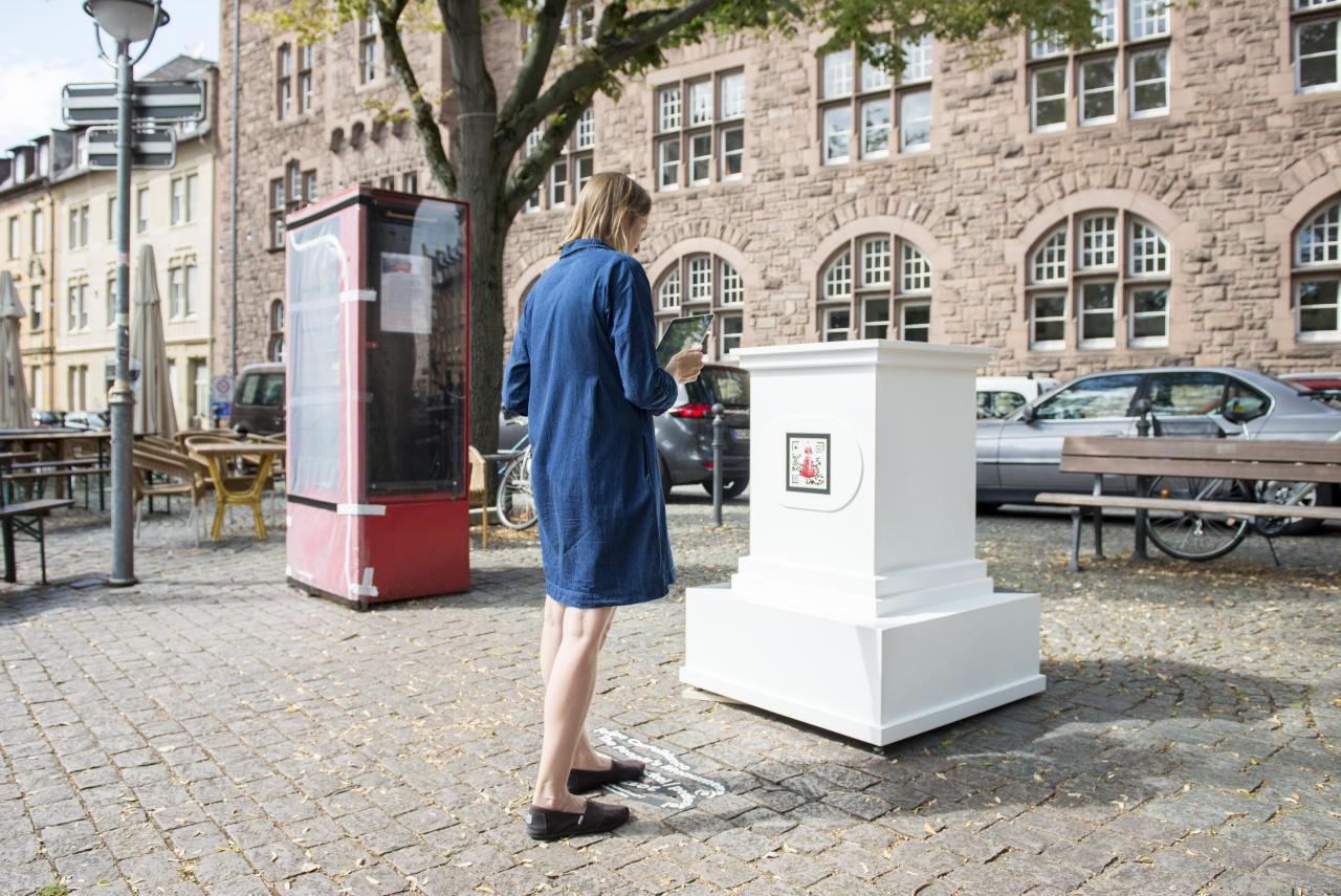 Eine Frau in blauem Kleid steht vor einem leeren weißen Sockel. Der Sockel gehört zum AR Kunstwerk »The People's Monuments«.