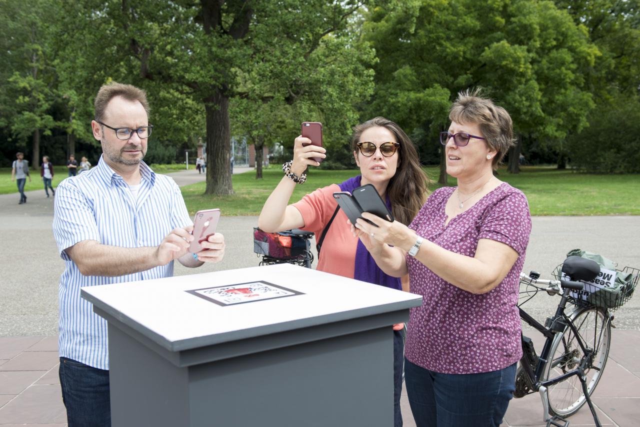 People with smartphones stand in front of an outdoor pedestal.