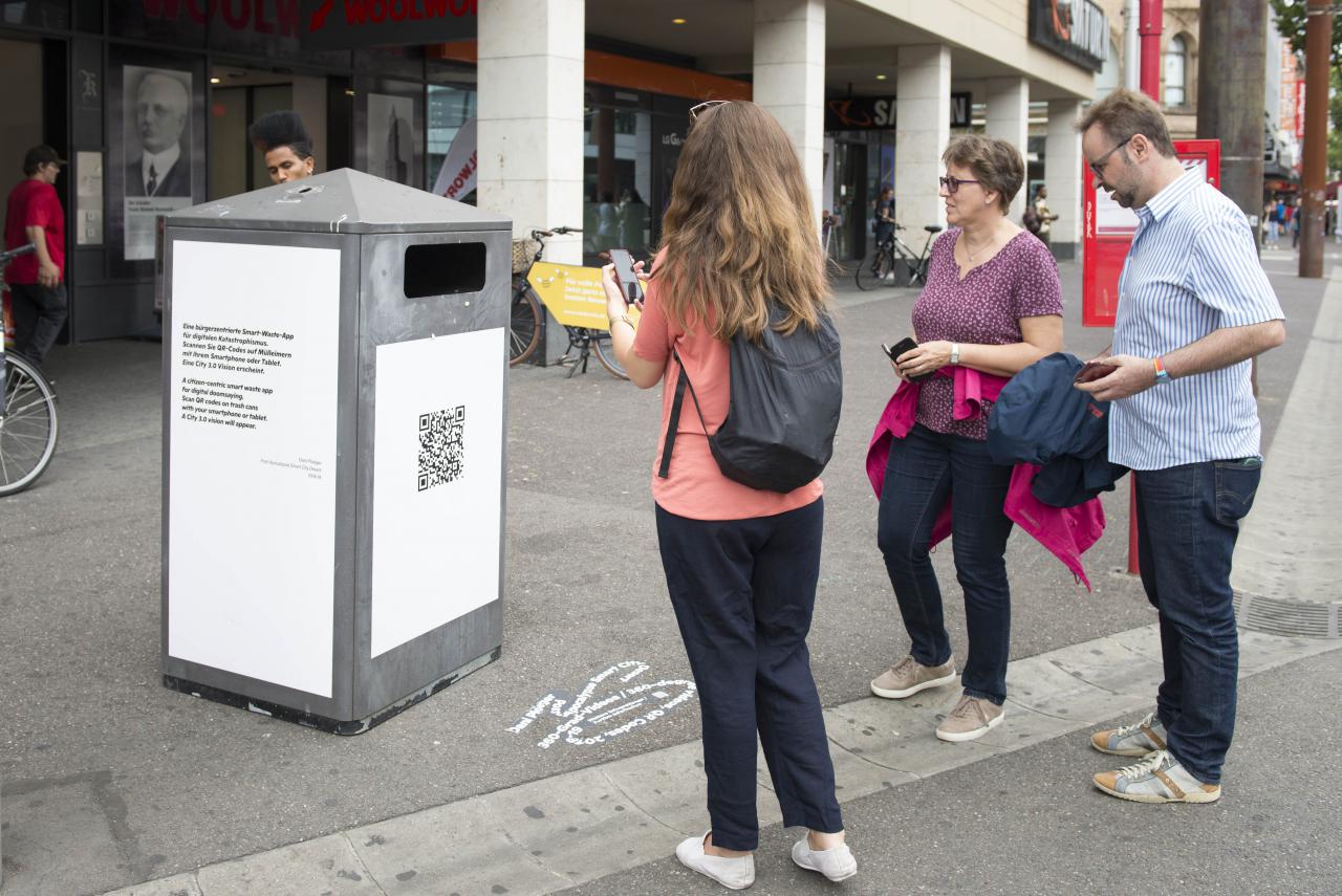 People with smartphones stand in front of a trash can with QR code
