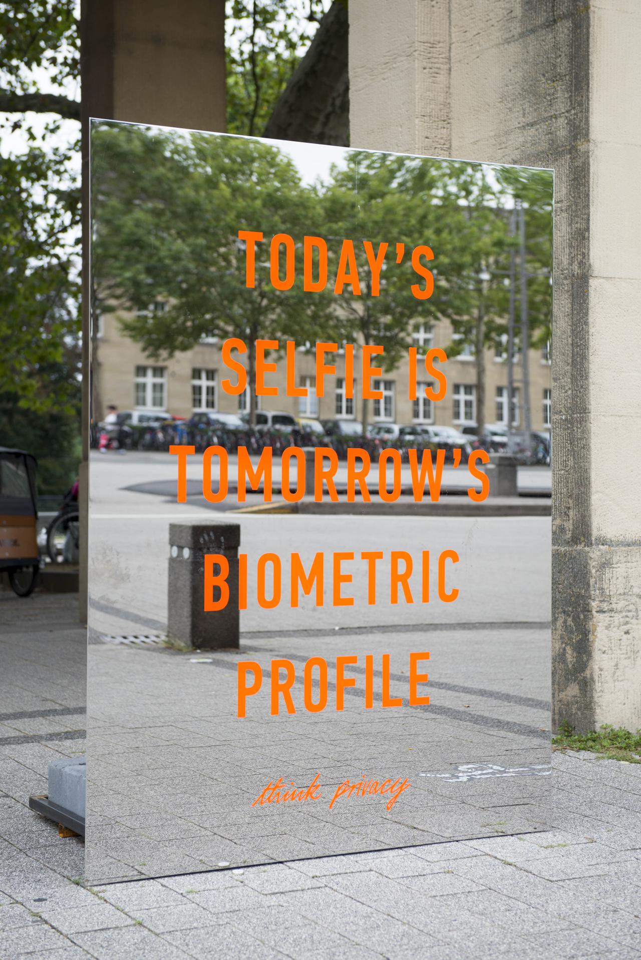 Zu sehen ist ein orangener Schriftzug »Todays Selfie is Tomorrows Biometric Profile« ist auf einem großen rechteckigen, rahmenlosen Spiegel. Die Installation steht draußen vor dem Eingang-Süd des Zoos.