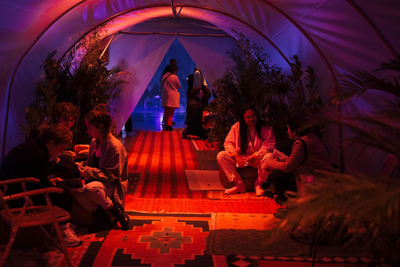 Here you can see the work »Sedekah Benih«. There are several people to see, who sit on carpets in an elongated tent. On the left and right are plants. The tent is illuminated with red light.