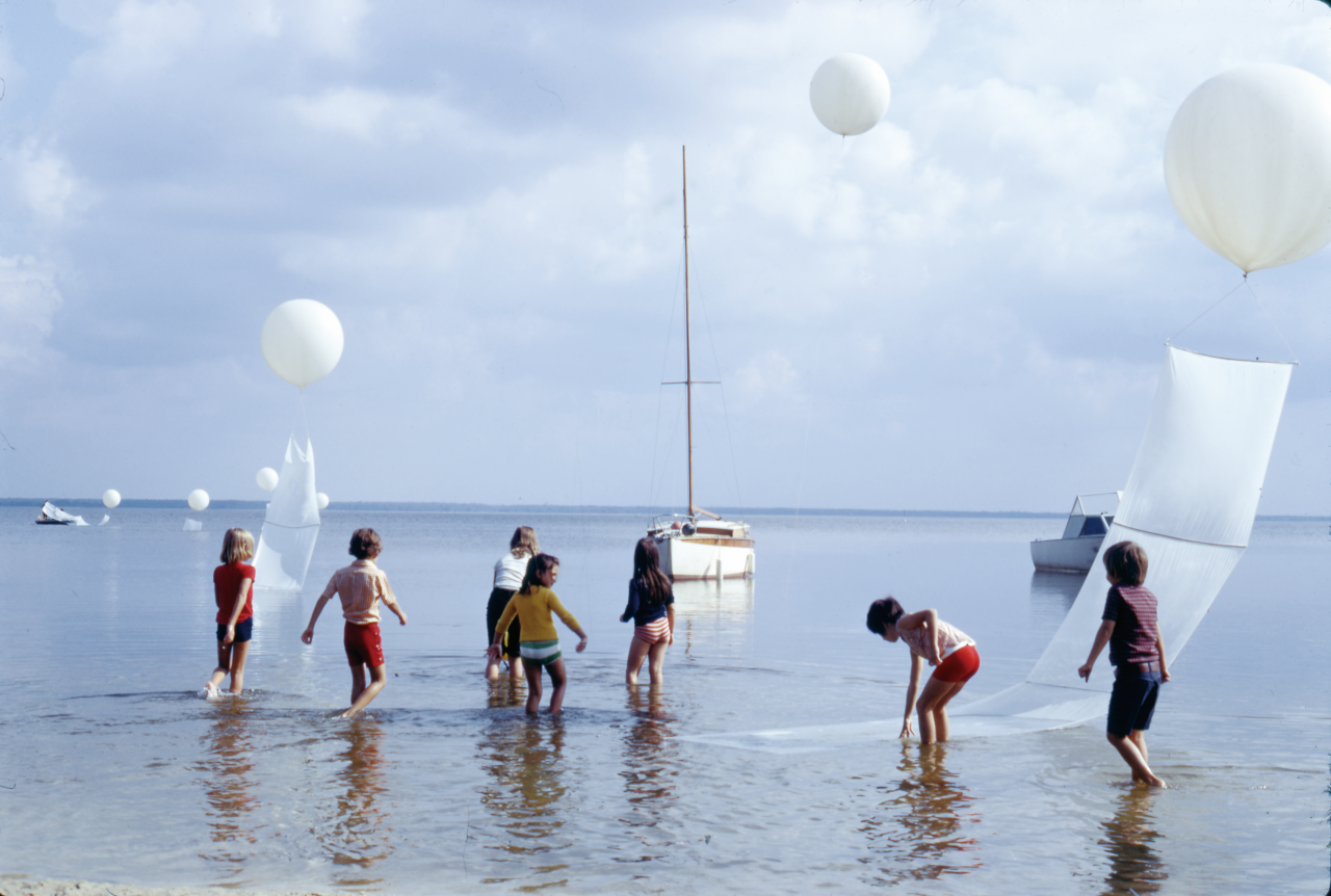 You can see several children running through water. Above them are white balloons. In the background you can see boats. 