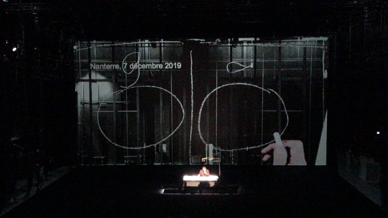 Bruno Latour sitzt auf einer Bühne hinter einem Schreibtisch. Hinter ihm ist eine große Leinwand, die zwei große Kreise zeigt, zwischen ihnen ist ein langer, senkrechter Strich.