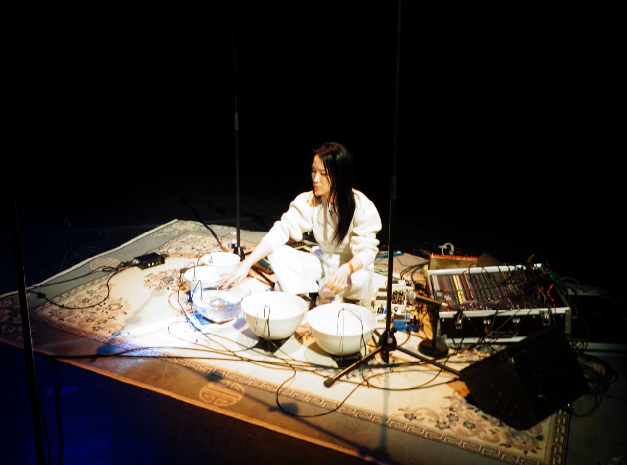 Tomoko Sauvage sitzt auf dem Boden, vor ihr stehen mit Wasser gefüllte Schalen und Mikrofone.