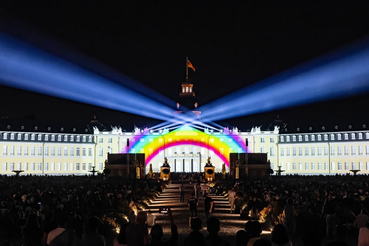 Zu sehen ist die Fasade des Karlsruher Schlosses. Darauf projeziert ist ein Regenbogen mit Scheinwerfern, die in den schwarzen Himmel leuchten.