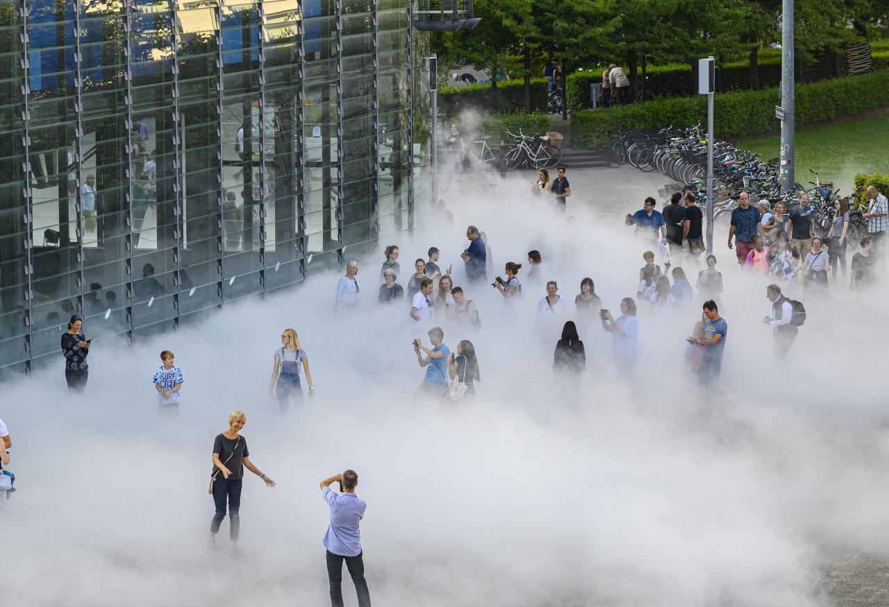 The photo shows a section of the glass cube of the ZKM in front of whose front right corner several visitors stand in the fog sculpture and take photos.