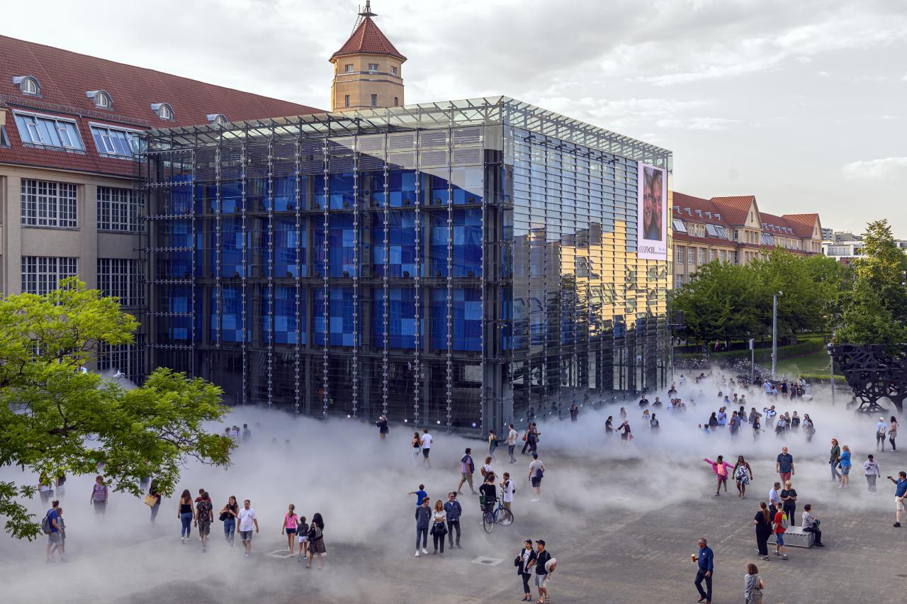 The photo was taken with a drone and shows the cube of the ZKM by day. A lot of people are gathered around the cube and are partly hidden behind the fog sculpture.