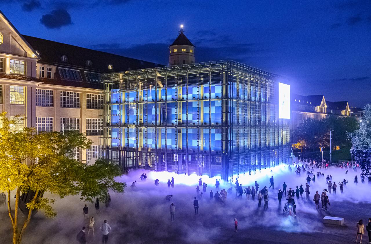 Das Foto ist bei Nacht mit einer Drohne aufgenommen. Es zeigt die Nebelskulptur und viele Silhouetten von BesucherInnen der KAMUNA 2019. Der Kubus und die Fassade des Gebäudes sind hell erleuchtet.  