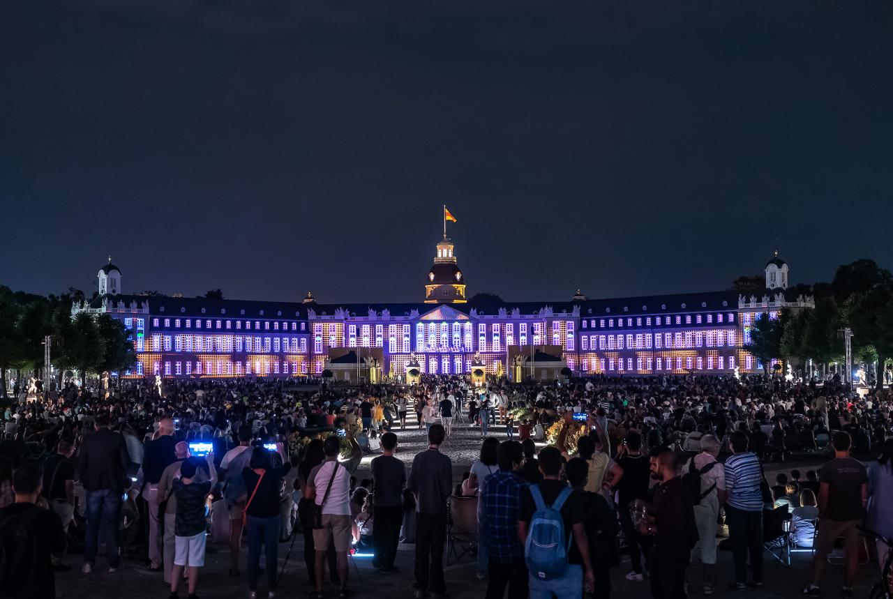 The black sky as a background highlights the brightly lit castle. Karlsruhe Castle illuminates in various shades of purple and pink, exploiting the structures of the castle to create geometric patterns.