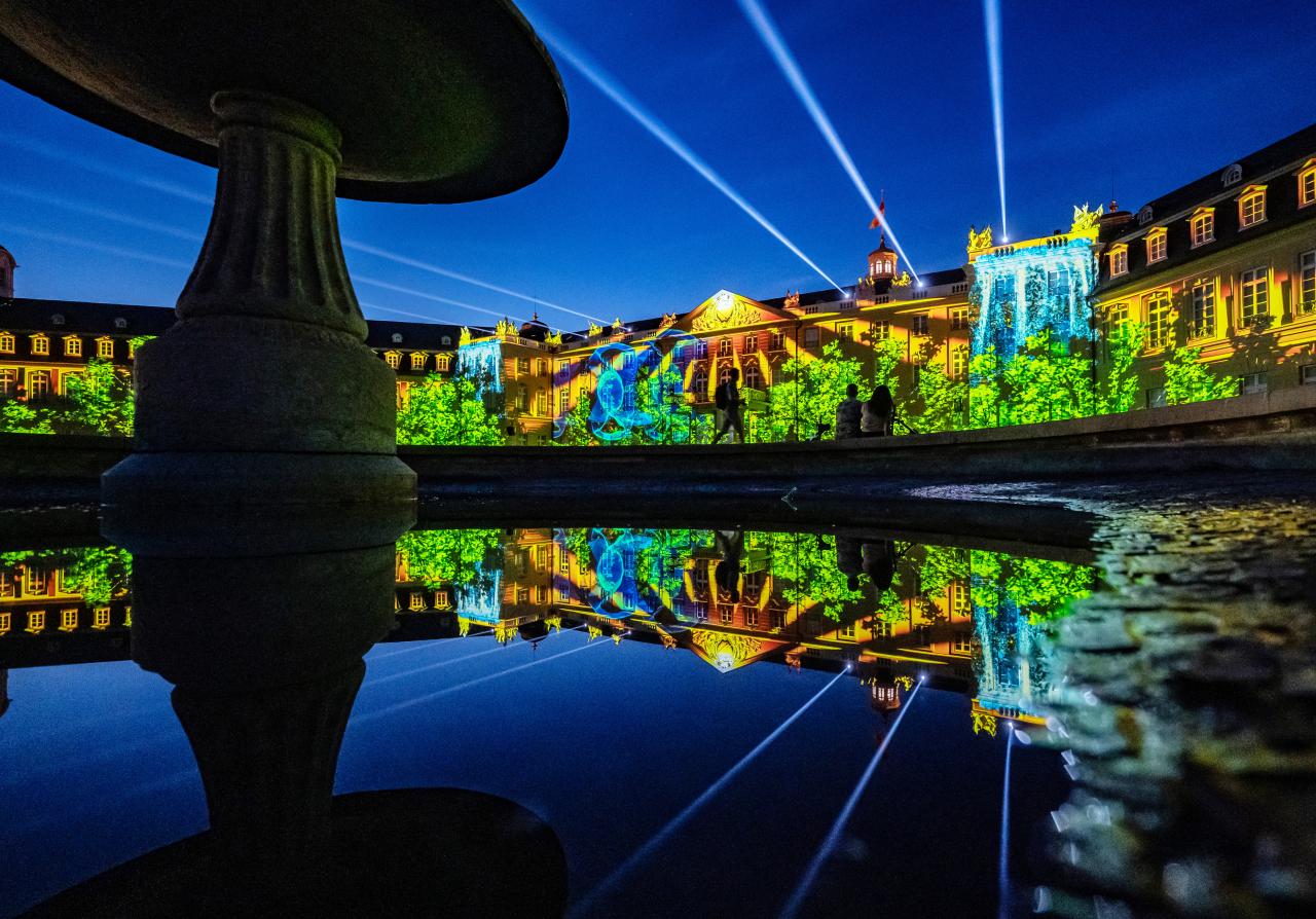 In the background you can see the illuminated facade of Karlsruhe Castle. A blue sky is projected over a landscape of trees with a waterfall. In the foreground the projection is reflected in a fountain. Spotlights rise into the sky.