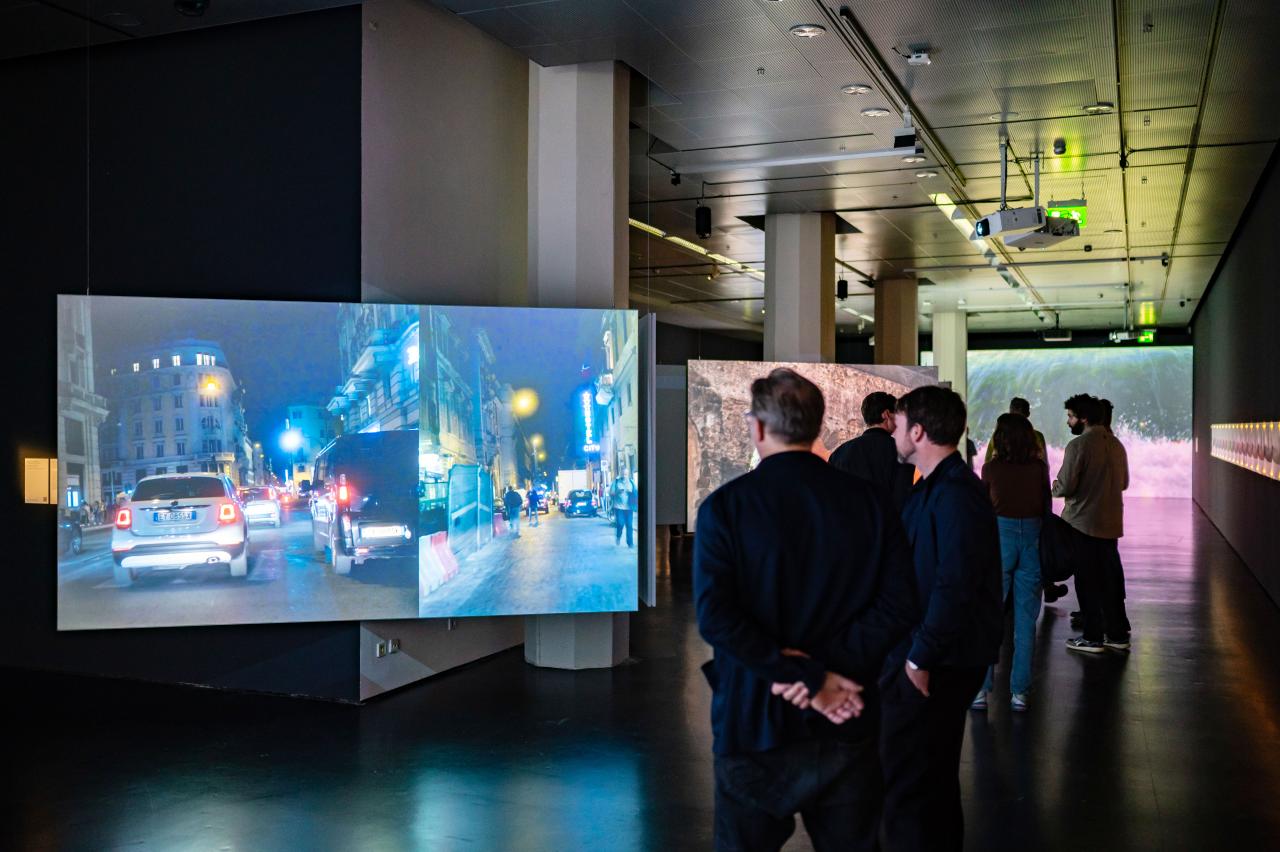 Zu sehen sind zwei Personen vor einem Bildschirm, auf dem ein Auto auf der Straße gezeigt wird.Ausstellungsansicht »Marijke van Warmerdam. Then, now, and then«.
