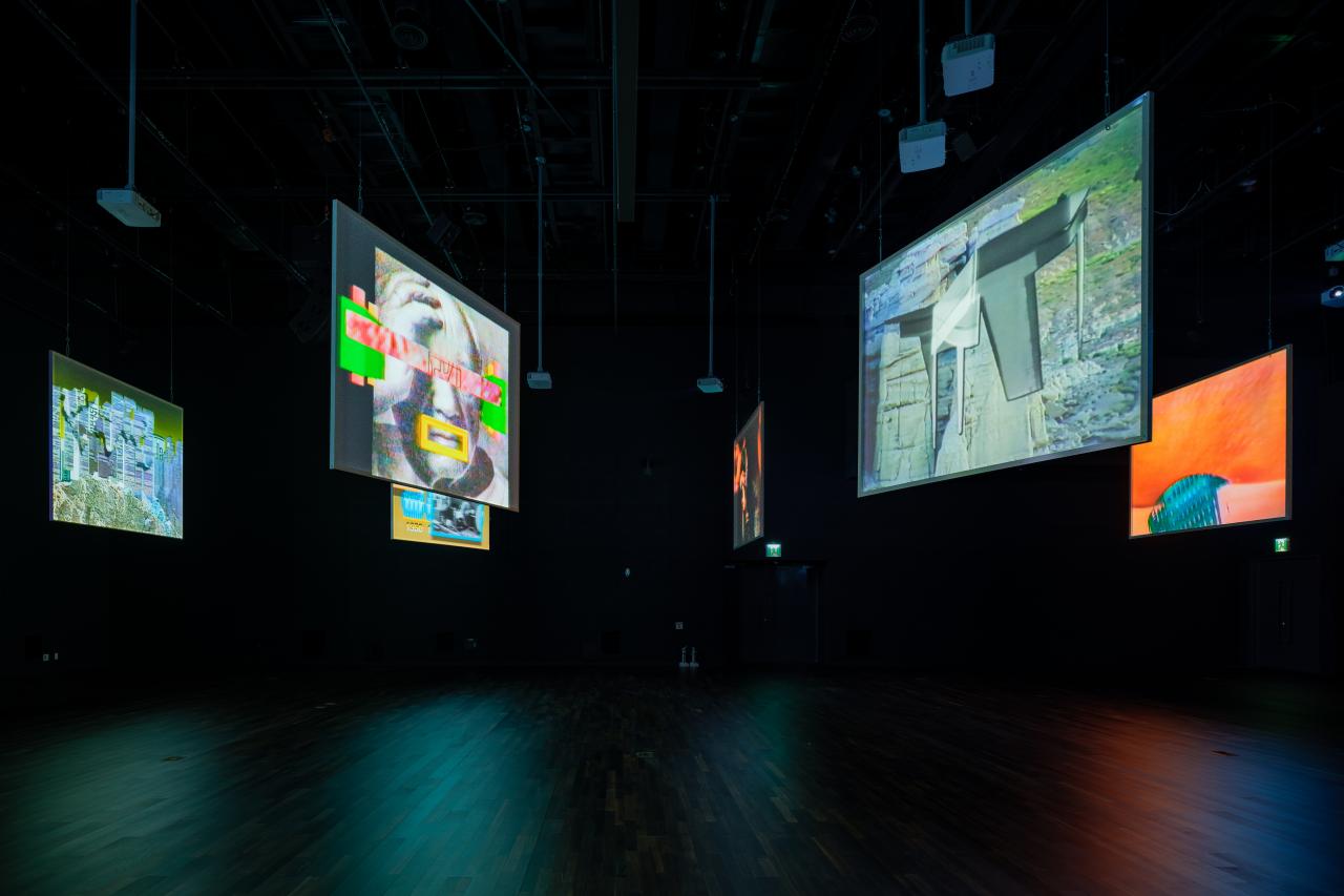 You can see several screens hanging from the ceiling in a darkened room. They show color-intensive video recordings.
