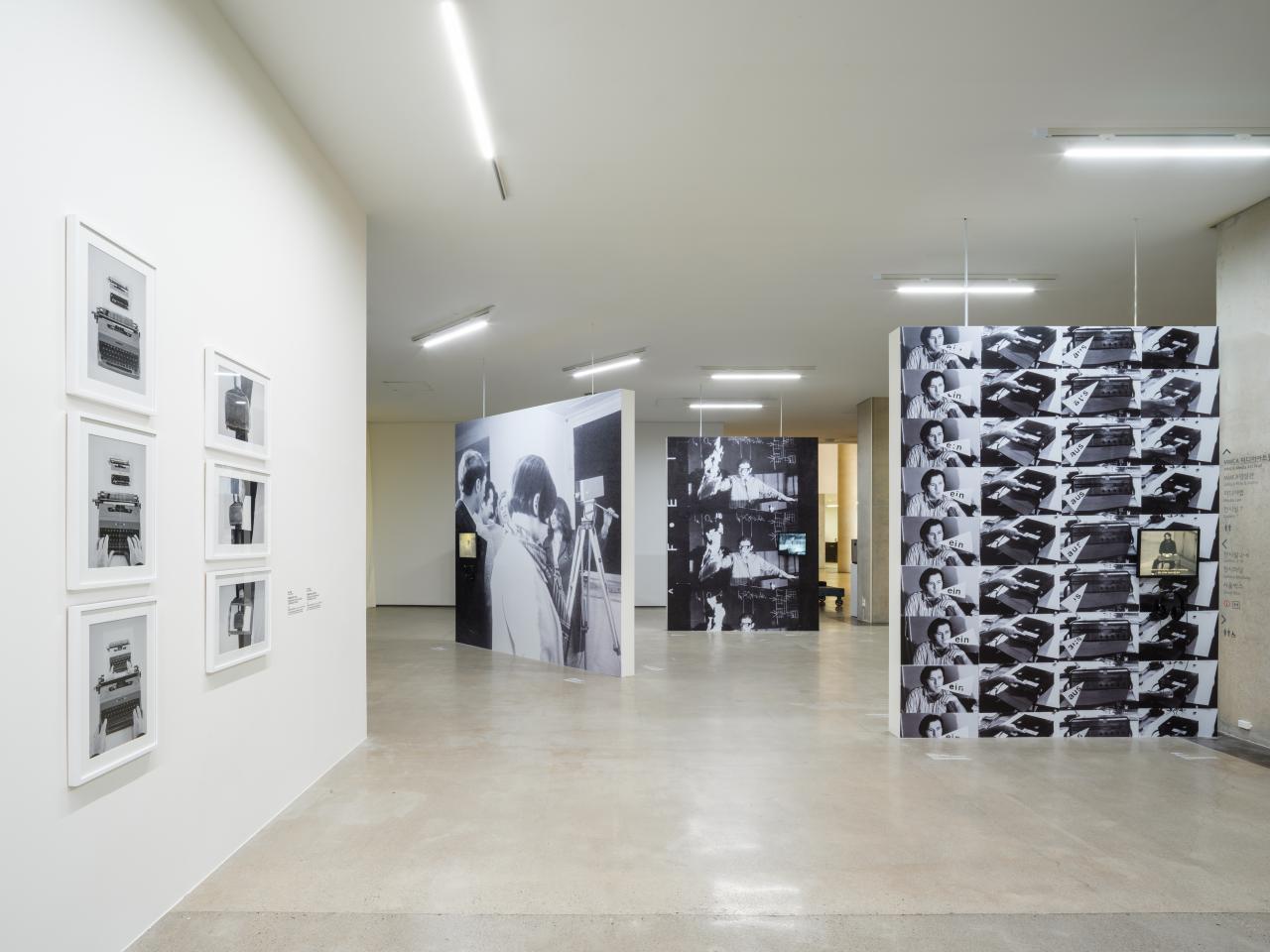 You can see an exhibition space. Three large black and white pictures divide the room. On the left wall hang 6 black and white photographs in white picture frames.