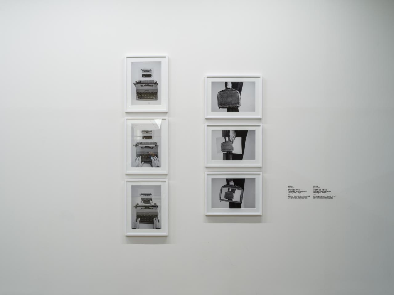 On view are six picture-in-picture, black and white photographs in picture frames on a wall. Three of them show a typewriter, the other three show a hand holding a briefcase.