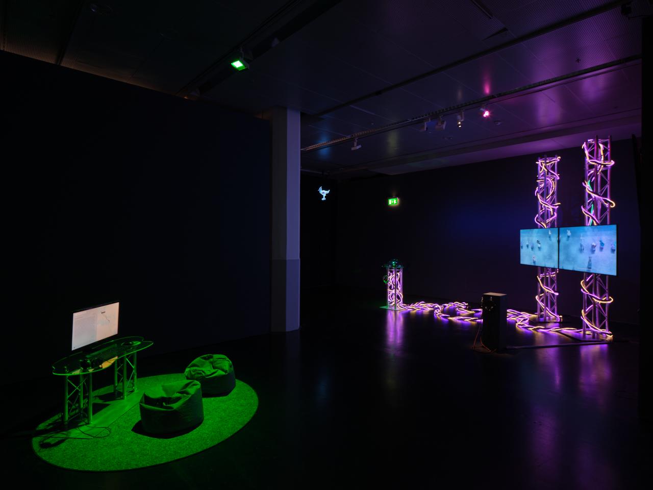 The picture shows a dark room with two works of art in the middle. On the left is a green-lit space with a computer and two beanbags and on the right is an installation with two screens surrounded by light elements. 