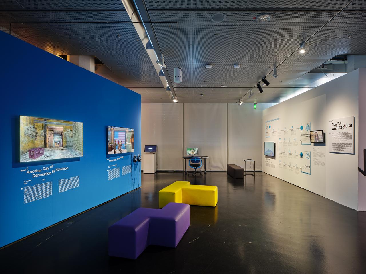 Blaue Wand in der Ausstellung »Playful Architectures« auf der linken Seite. Daran angebracht sind zwei Bildschirme, die Computerspiel zeigen. Im restlichen Ausstellungsraum verteilen sich weitere Bildschirme.