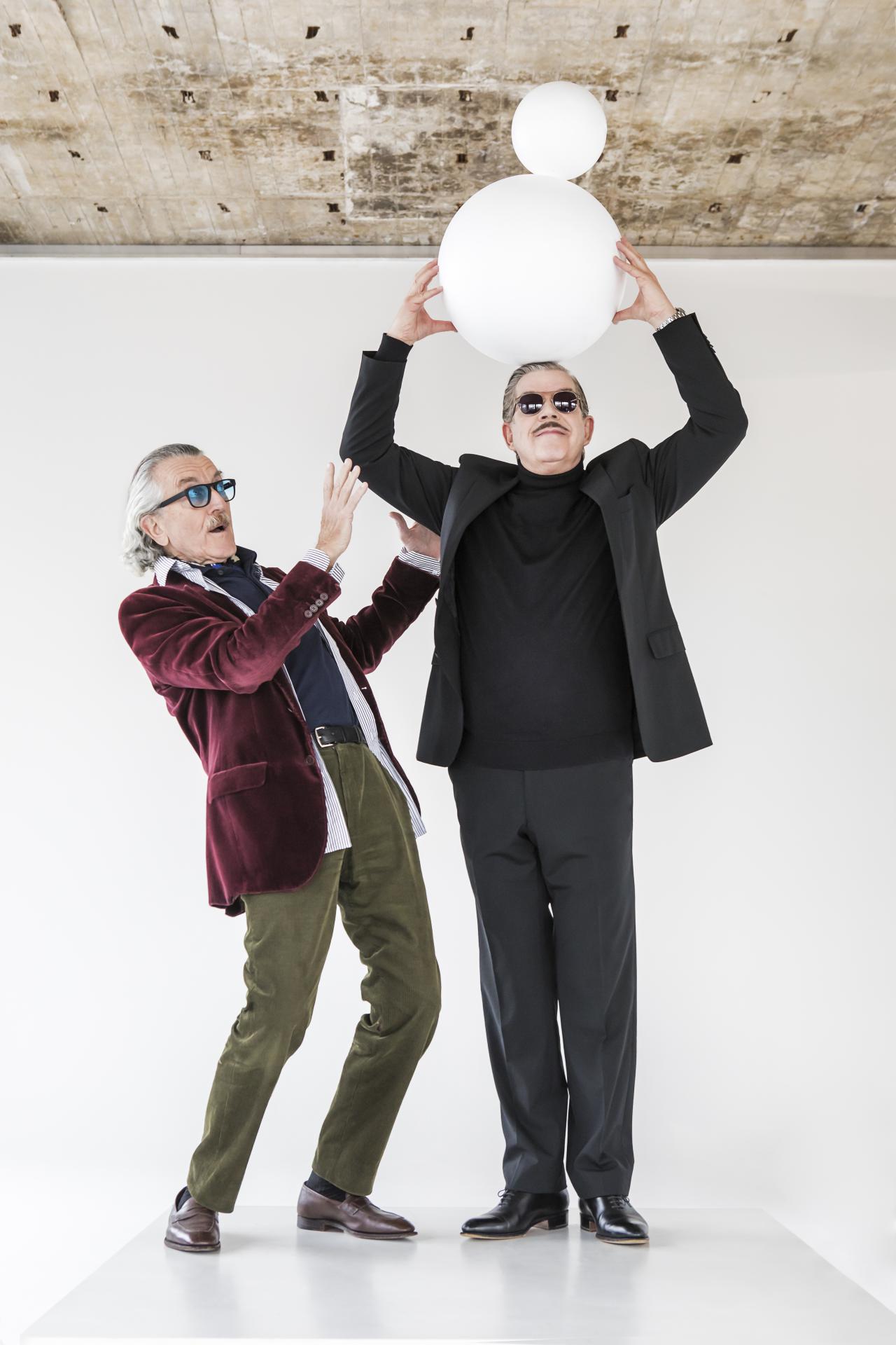 The musician duo Yello stands side by side in front of a wall. The one on the right wears a suit and holds a sculpture of two round balls above his head. The left one stands next to him in an intercepting position.