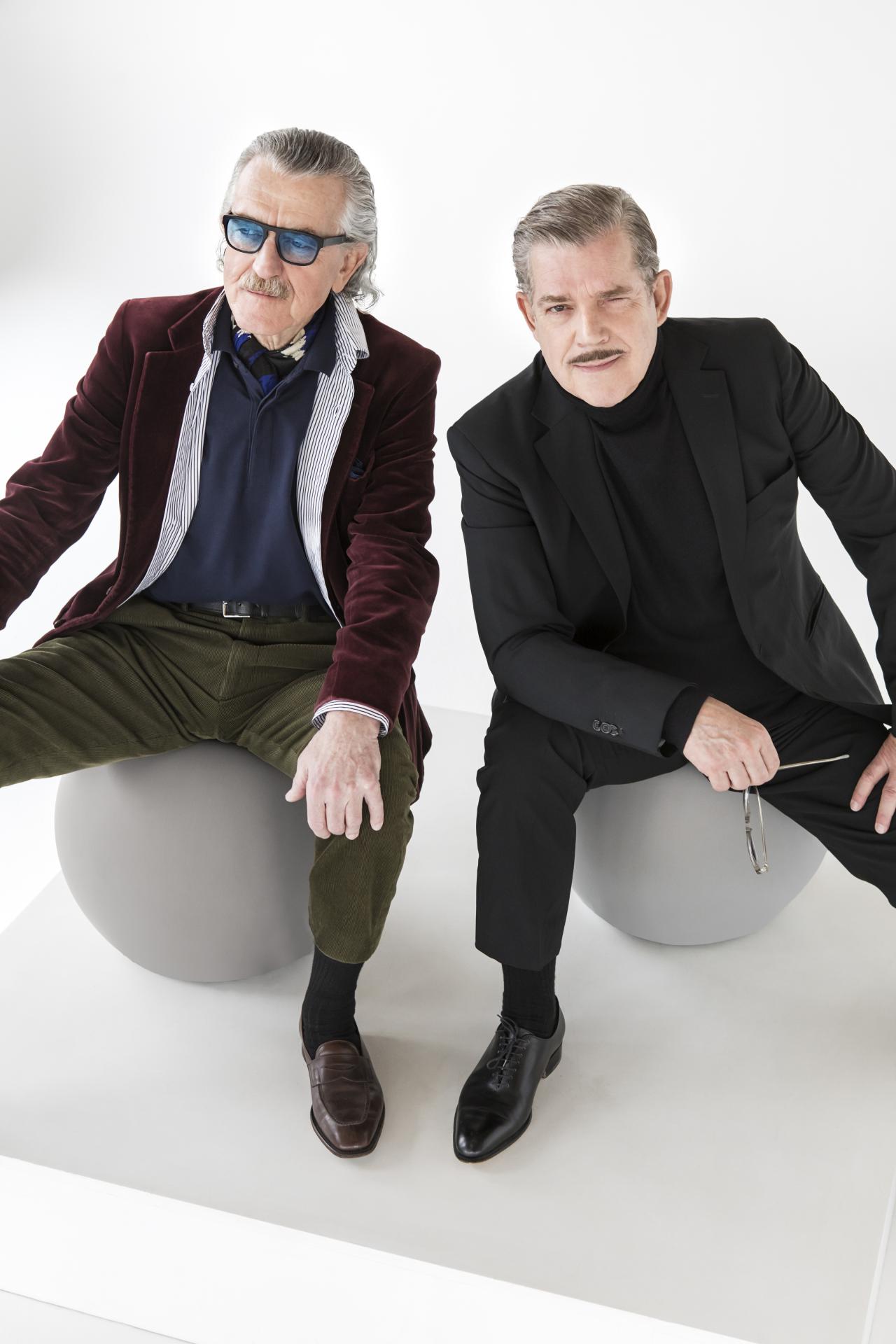 Two men sit next to each other on one ball. They both wear casual suits. The left one wears colored glasses, both wear moustaches.