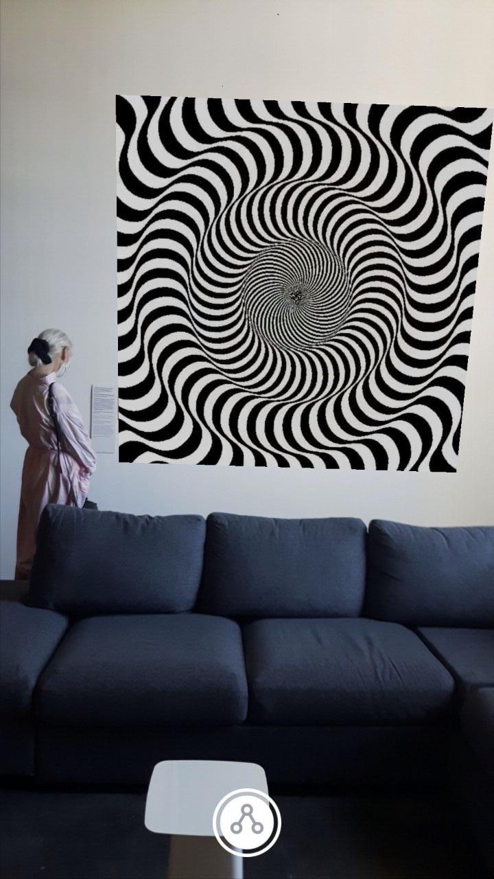 A woman stands behind a sofa, in front of a picture. The picture shows an optical illusion of black and white lines that simulate movement to the eye.