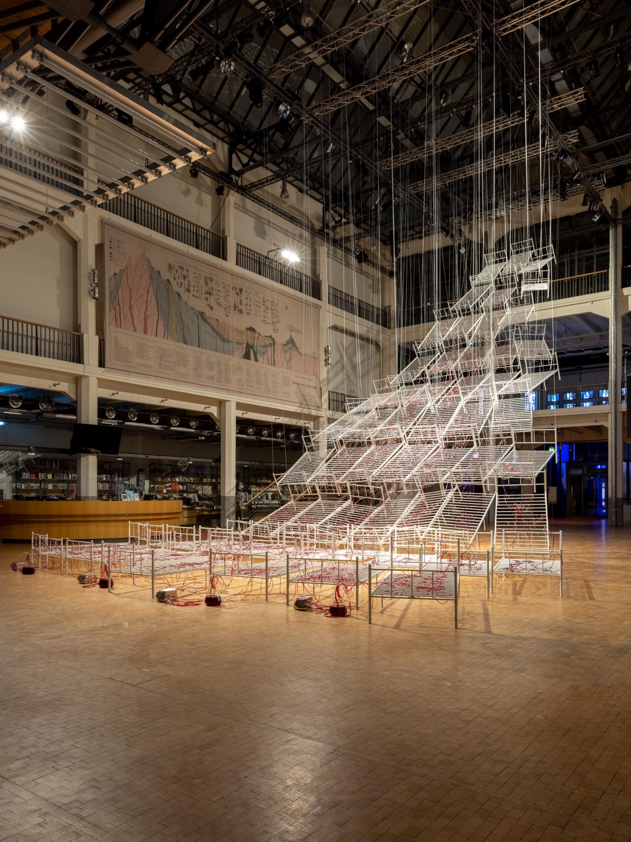 Installation view of Chiaru Shiota's »Connected to Life«. Hospital bed frames can be seen hanging from the ceiling in the shape of a pyramid. These are crisscrossed by tubes through which red paint flows.