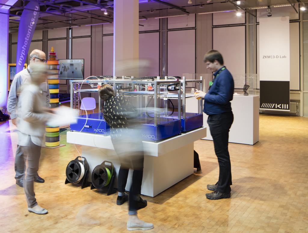 Vier 3D-Drucker vor denen Menschen stehen und zuschauen