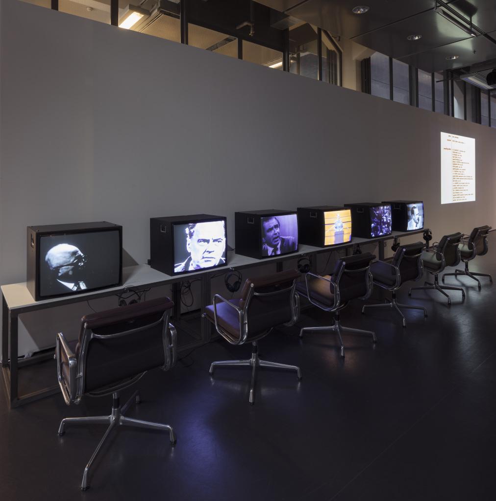 An einer Wand, ein langer Tisch mit sechs Fernsehern, auf denen Unterschiedliche Videos laufen. Dazu jeweils Kopfhörer und davor schwarze Stühle.