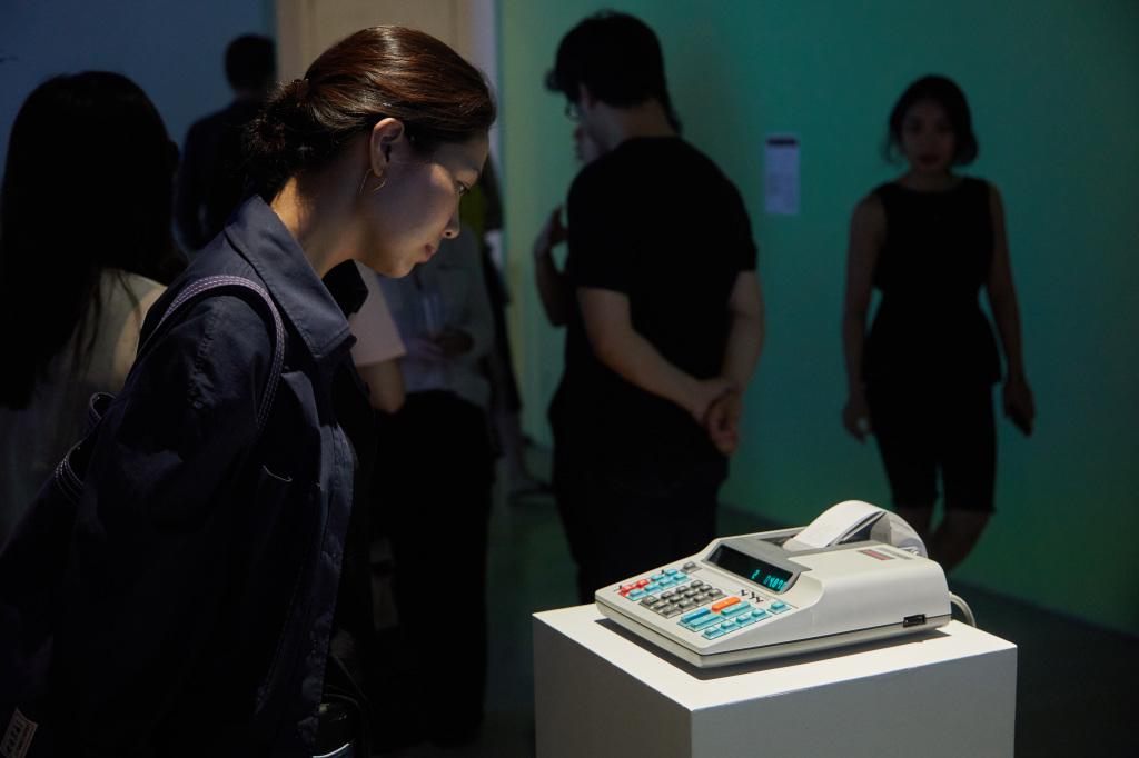 Foto der Ausstellung »Open Codes. Connected Bots«. Ein alter Taschenrechner mit Thermopapier-Rolle rechnet um Bitcoins zu generieren.