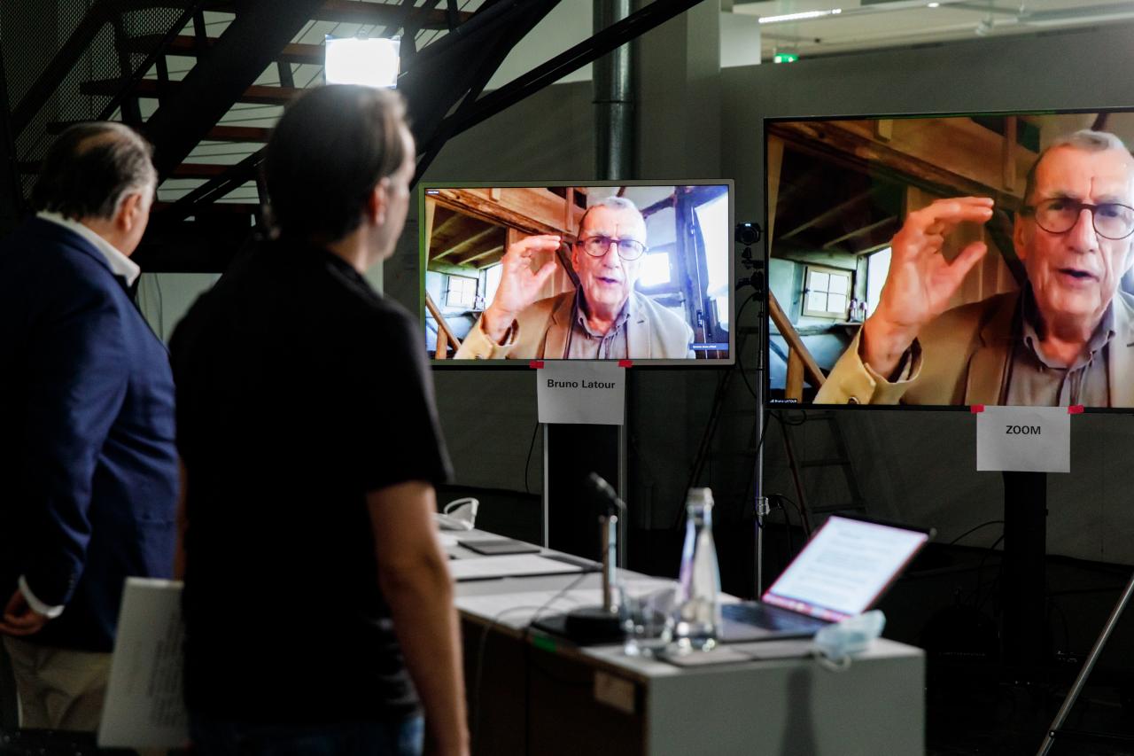 Bruno Latour ist in zwei Bildschirmen zu sehen. Zwei Männer, einer davon Peter Weibel, stehen vor den beiden Monitoren.