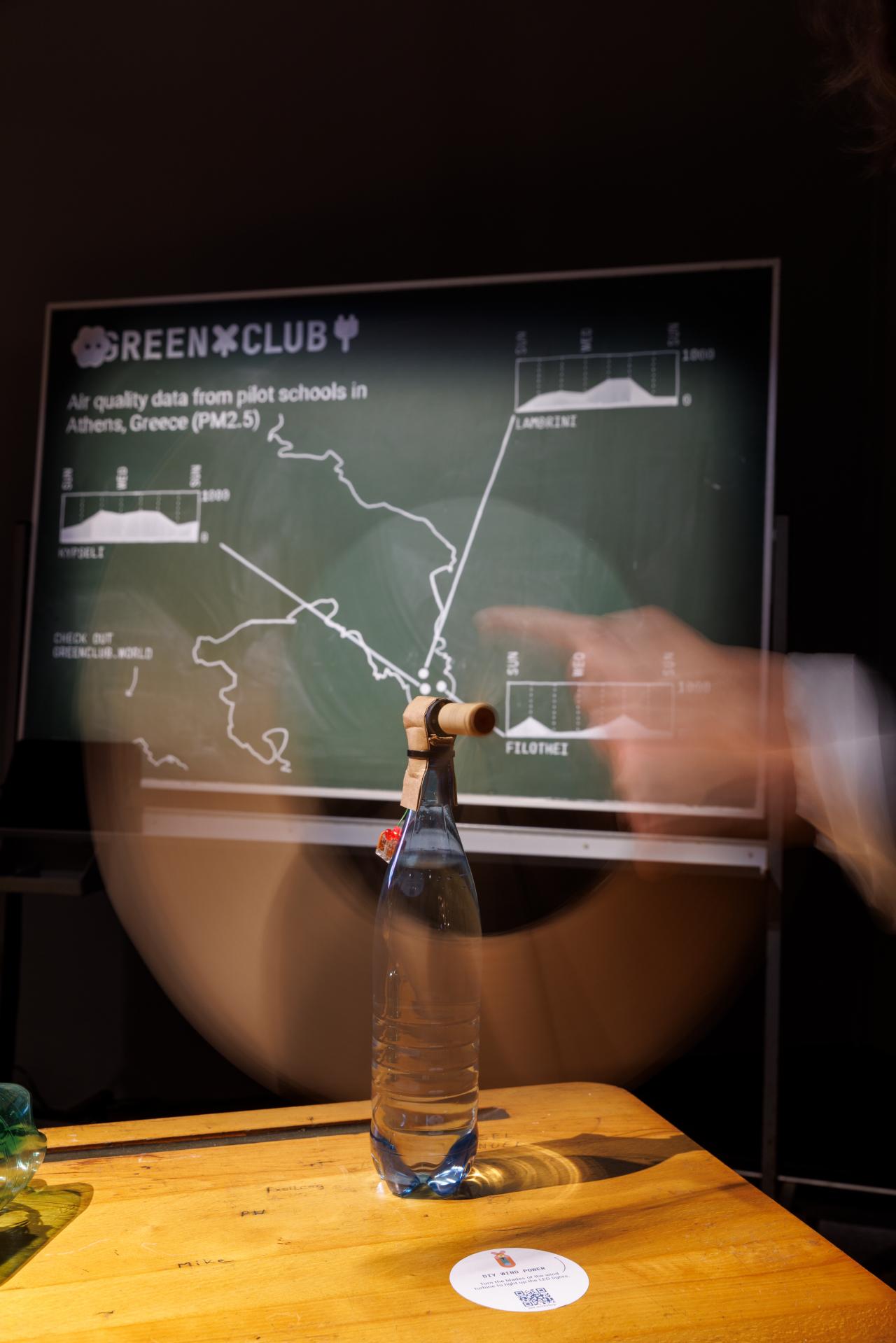 Im Vordergrund steht eine Glasflasche, auf der eine Art Rohr befestigt wurde. Im Hintergrund ist eine Tafel mit einer Statistik zu sehen.