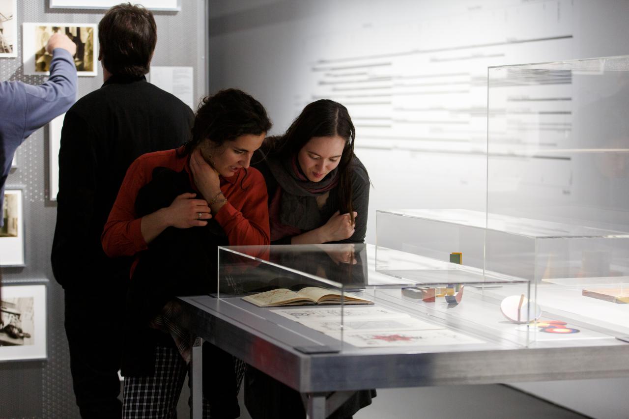On view are two women who bend over a display case in the exhibition and take a closer look at the book on display.