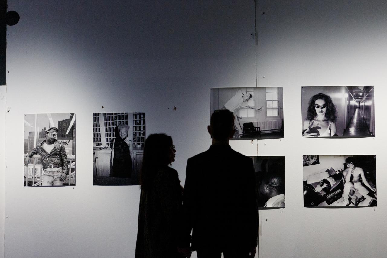Zwei Personen, die nur als Schatten zu erkennen sind, stehen vor einer Wand mit mehreren Schwarz-Weiß-Fotografien.