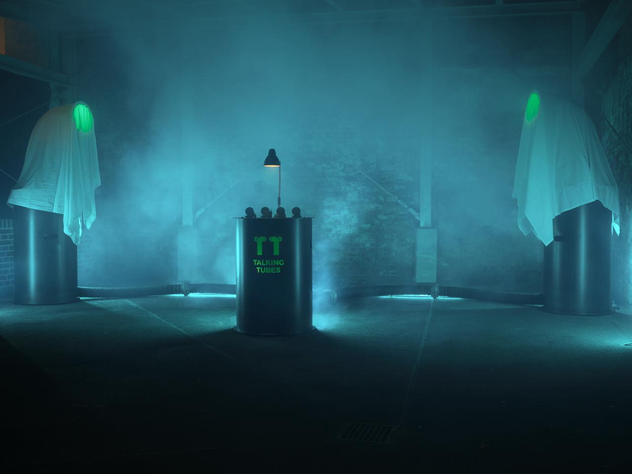 A blue-lit, foggy room with the Talking Tubes installation: a control panel between two large tubes covered by white blankets