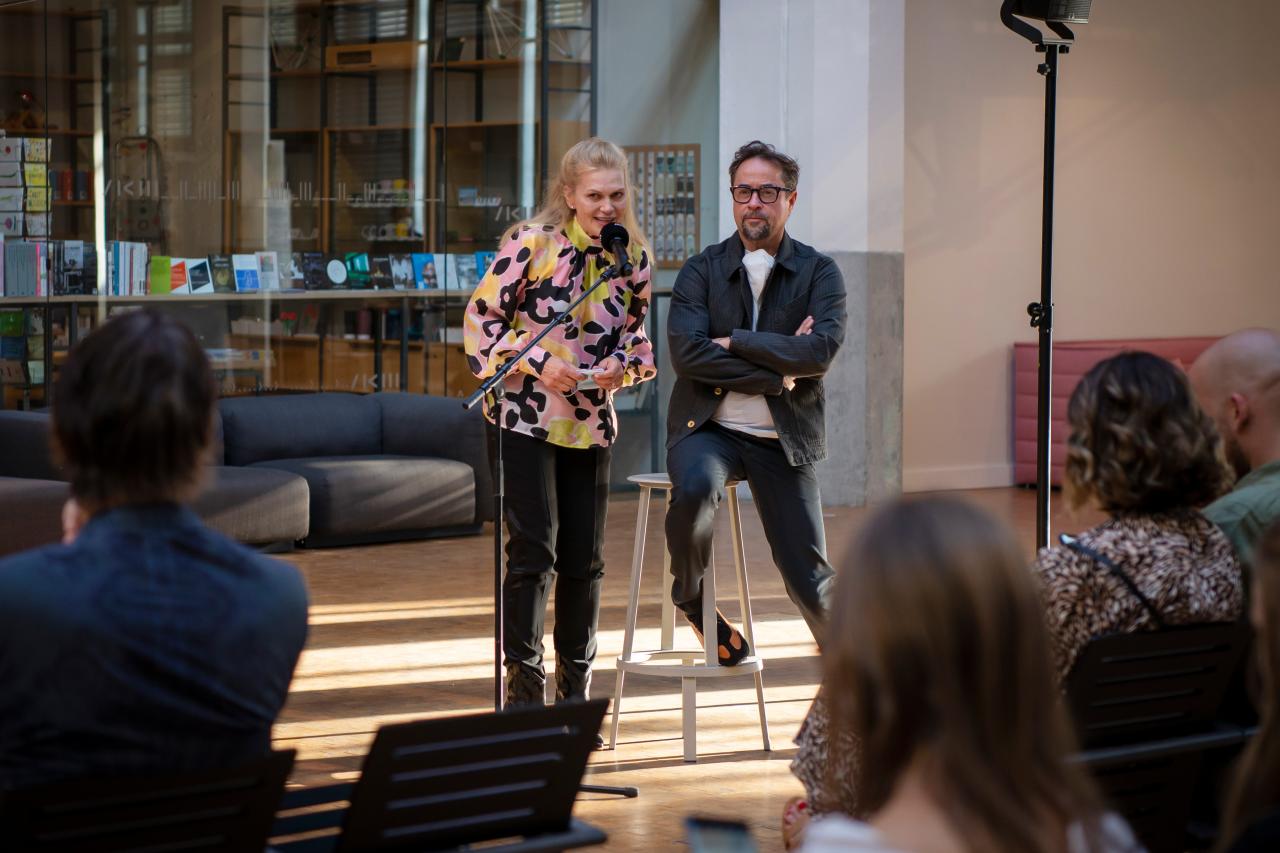 The actors Anna Loos and Jan Josef Liefers are behind a microphone. Anna Loos speaks, Jan Josef Liefers sits on a stool, arms folded, and listens.