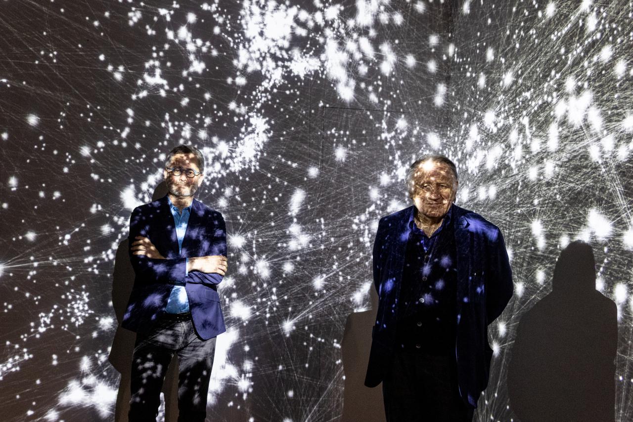Zwei Männer stehen im Ausstellungsraum vor einer Projektion, die den Kosmos als Netzwerk zeigt. Sie sind von der Projektion überdeckt. Der linke Mann ist der Wissenschaftler Albert-Lázsló Barabási. Der rechte Mann ist Peter Weibel. Beide lächeln.
