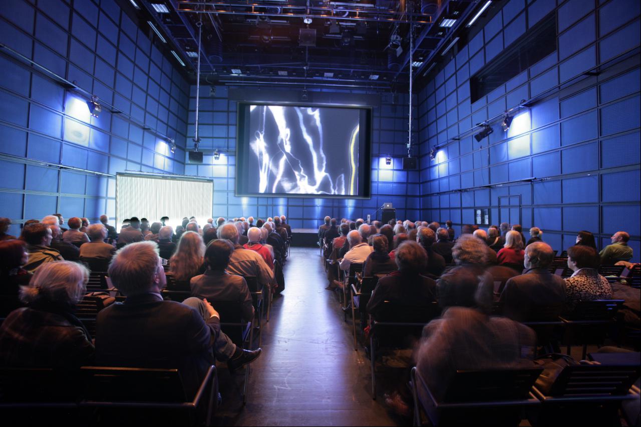 Das ZKM_Medientheater voller Menschen, die auf eine Leinwand schauen, auf der amorphe Strukturen in weiß zu sehen sind