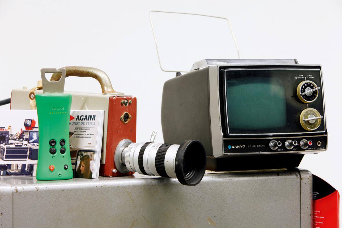 Ein sehr alter, kleiner TV-Monitor, eine alte Videokamera und eine Fernbedienung mit integriertem Flaschenöffner 