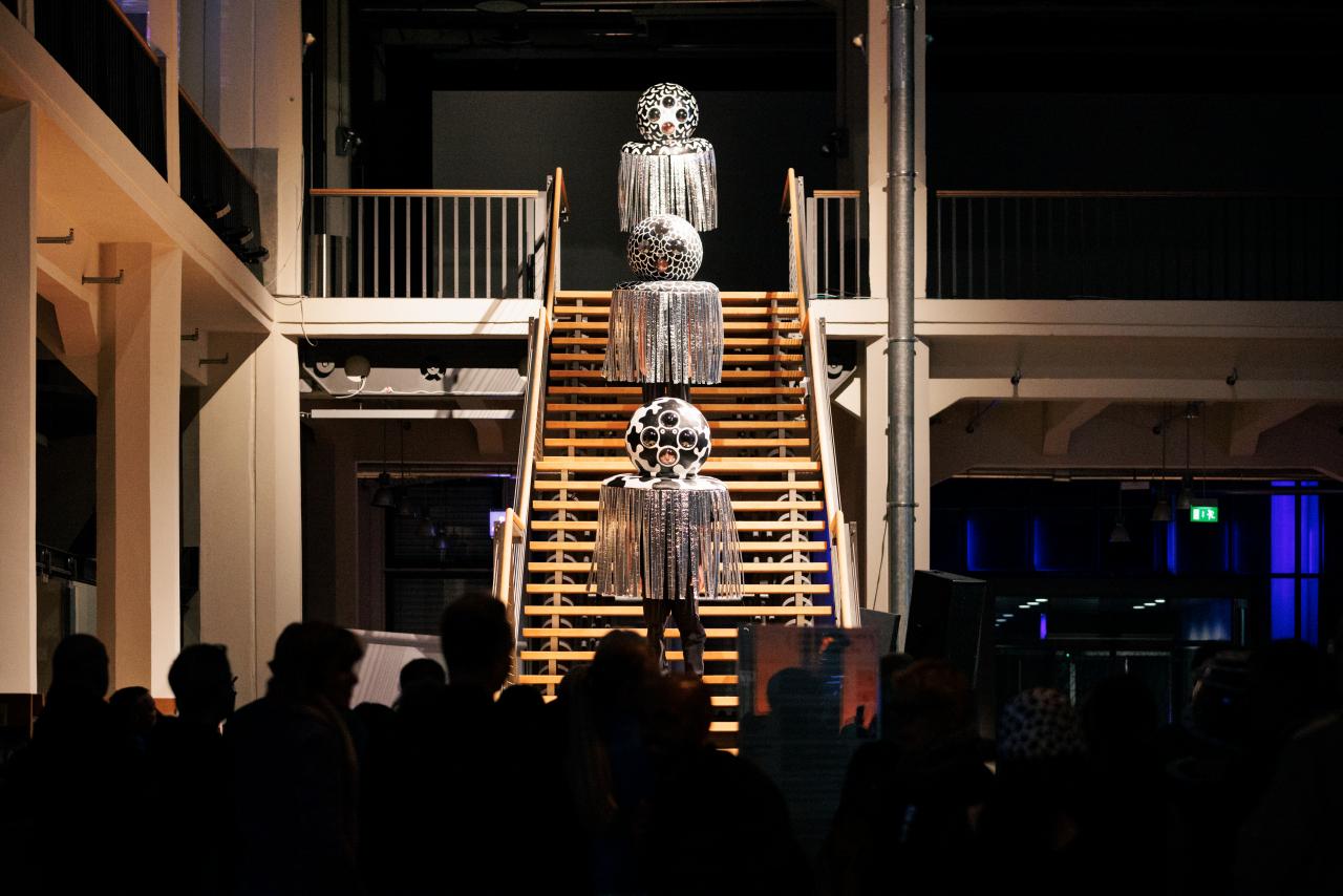 A performance is running on the stairs in the foyer of the ZKM. At intervals of a few steps, three people stand on the stairs and carry a ball above their heads with strings hanging down.