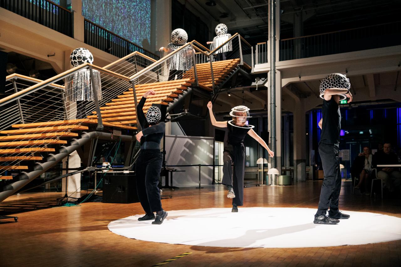 Fünf verkleidete Menschen tanzen ihre performance im Foyer des ZKM. Ihre Verkleidung besteht aus einer geschmückten Kugel über ihrem Kopf, sonst sind sie schwarz angeszogen.