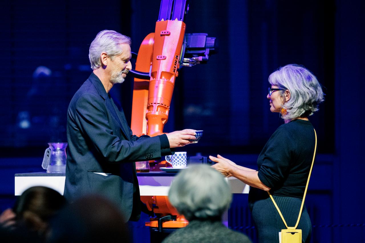 Im Hintergrund ist ein orangener Roboter, der gerade tee gemacht hat, den der künstlerisch-wissenschaftliche Leiter des ZKM | Karlsruhe Alistair Hudson einer Frau überreicht.