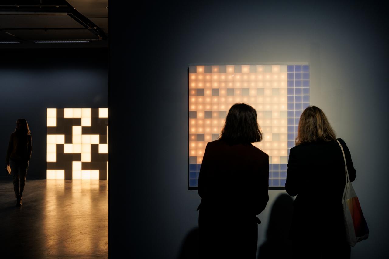 Two people are seen in front of a luminous Walter Giers work. In the background is another cube-shaped luminous object.