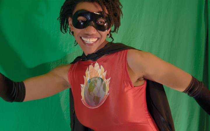 Eine Frau hat ein Superhelden-Kostüm an, lächelt und ballt ihre rechte Faust in die Höhe.