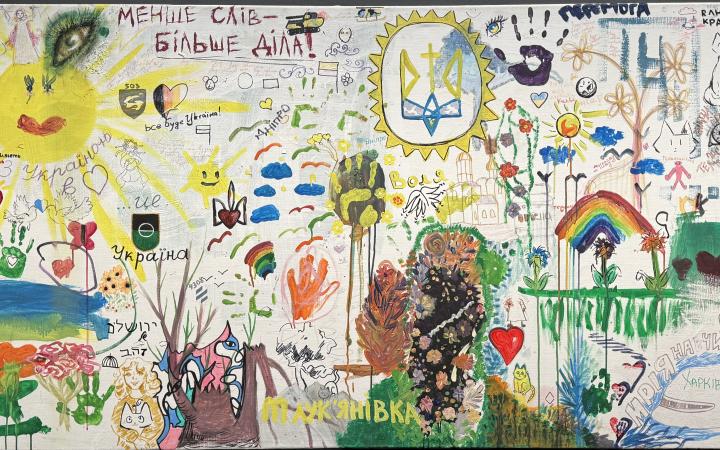The picture is full of colorful little creations. A yellow, laughing sun dominates on the left, many handprints and a colorful rainbow on the right, as well as flowers, people and hearts.