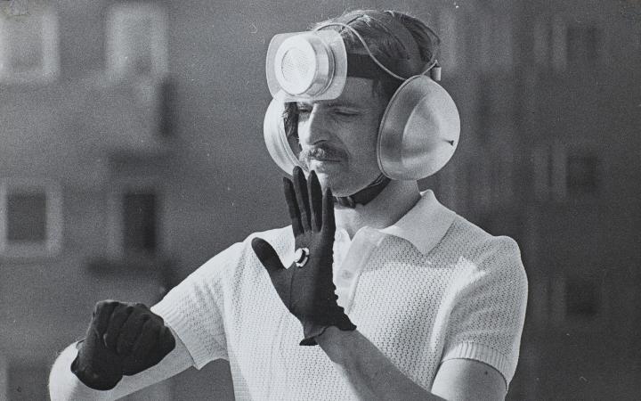 Das Schwarzweiß-Photo zeigt einen Mann mit einer technischen Installation auf dem Kopf, seine Ohren sind mit Schallschützern bedeckt.