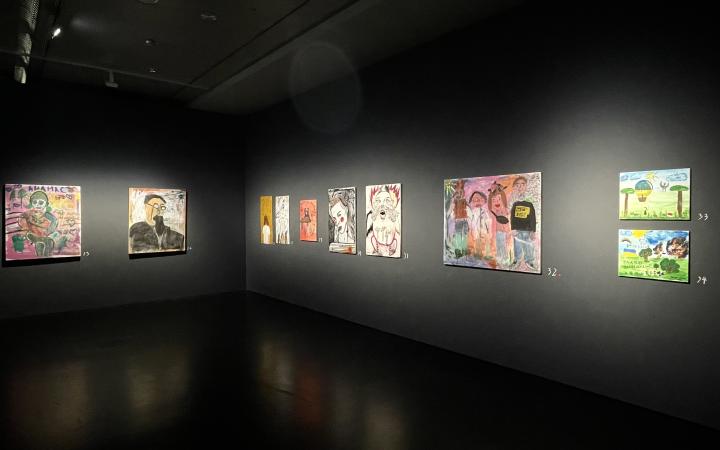 Ausstellungsansicht aus der Ausstellung "Wenn der Krieg ins Museum kommt". Zu ist ein Raum an dessen Wand viele der gemalten Bilder hängen und links ins eine Zone mit Schuhen und einem Computer, während rechts zwischen Holzkisten stehen