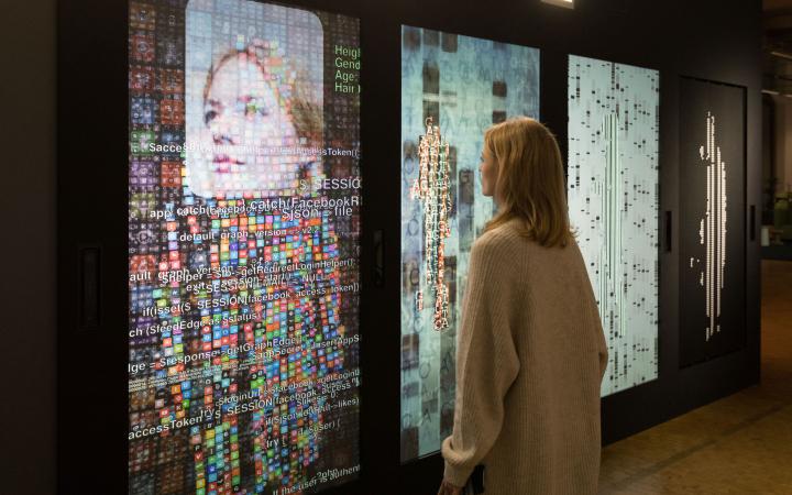 A woman stands in front of a wall with several screens and digital representations