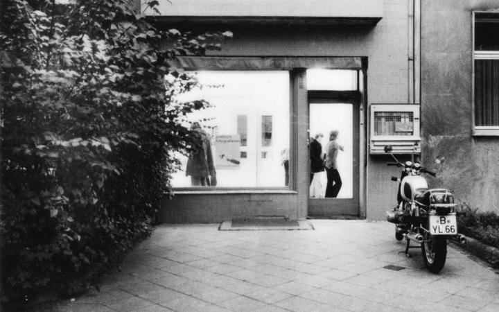 Black and white photo shows the street view of a gallery space. In front of it is a motorcycle.