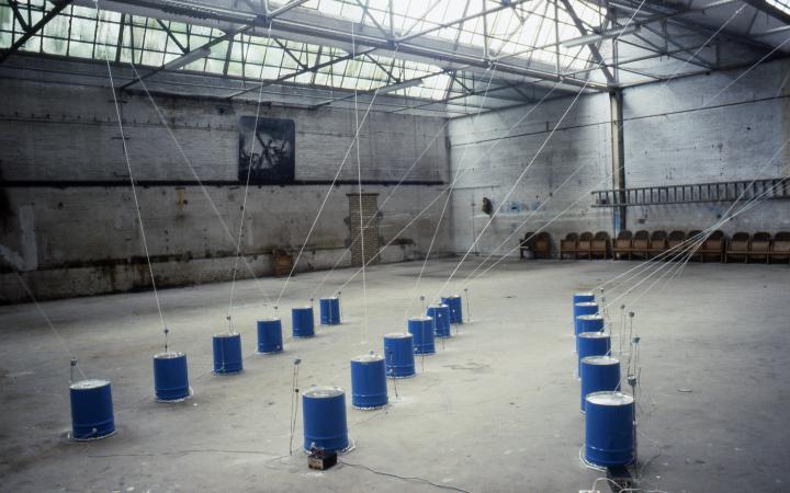 The picture shows an installation by Paul Panhuysen: Several dark blue barrels, from which ropes go to the ceiling, stand in a warehouse. 