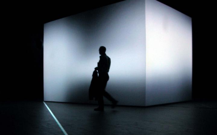 In einem völlig dunklen Raum steht ein 3,5 Meter hoher Würfel, dessen Form durch eine Lichtprojektion verfremdet wird. Davor der Schatten eines Mannes..